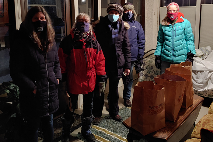 Five people in masks and heavy winter clothes inside a front porch with four brown paper grocery bags on a small table